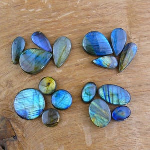 May include: Assortment of polished labradorite gemstones in various shapes, including teardrops and ovals. The stones display iridescent blue, green, and gold flashes, set against a wooden background. Ideal for jewelry making or collecting.