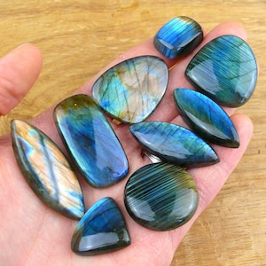 Può includere: Una collezione di pietre preziose in labradorite lucida di varie forme, tra cui ovali, rettangolari e triangolari. Ogni pietra presenta una superficie liscia e riflettente con bagliori iridescenti di luce blu, verde e dorata su base scura.