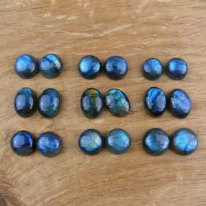 Könnte beinhalten: Eine Sammlung polierter Labradorit-Edelsteine in verschiedenen Formen, darunter ovale und runde Cabochons. Die Steine zeigen ein lebendiges Blau und schillernden Glanz, mit Andeutungen von Grün und Gold. Die Edelsteine liegen auf einer Holzoberfläche.