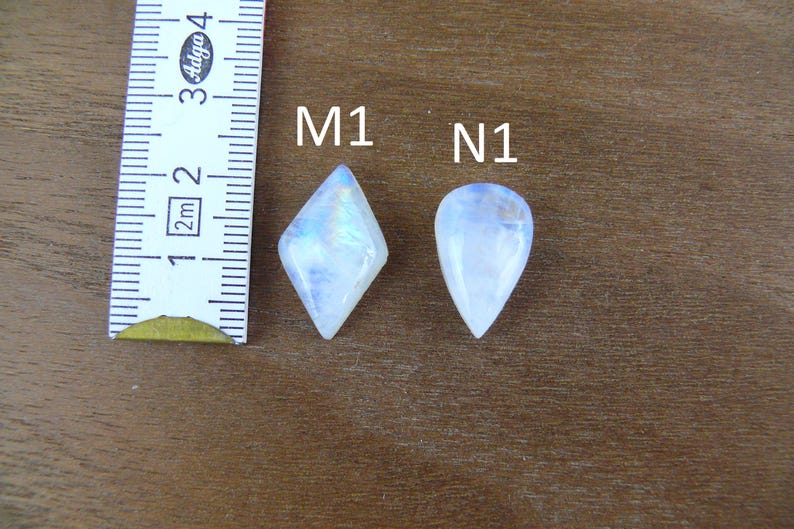 May include: Two moonstone cabochons labeled M1 and N1 next to a ruler. The M1 stone is diamond-shaped, and the N1 stone is pear-shaped. Both stones exhibit a translucent white appearance with blue iridescence. The ruler shows a scale in centimeters.