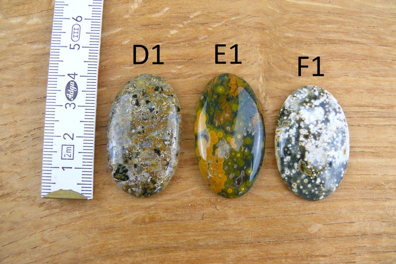 Op de afbeelding: Drie ovaalvormige gepolijste edelstenen met de labels D1, E1 en F1, geplaatst naast een liniaal met centimeters. De stenen hebben gevarieerde gespikkelde en gevlekte patronen in tinten groen, geel, bruin en wit.