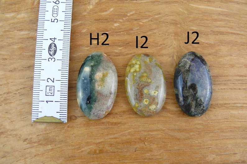 Op de afbeelding: Drie ovaalvormige gepolijste edelstenen met de labels H2, I2 en J2, geplaatst naast een witte liniaal met metingen in centimeters. De stenen vertonen gevarieerde patronen van groene, bruine, gele en donkergrijze insluitingen in het oppervlak.