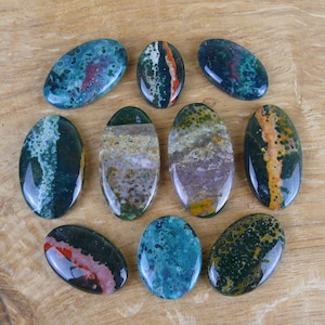 Puede incluir: Diez piedras pulidas de forma ovalada con una variedad de colores y patrones, incluyendo verde, azul, rojo, naranja y marrón. Las piedras están dispuestas en círculo sobre una superficie de madera.