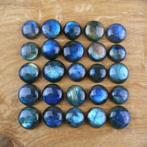 Könnte beinhalten: Vierundzwanzig runde Labradorit-Cabochons mit irisierenden blauen, grünen und goldenen Reflexen. Die polierten Steine sind auf einer hellbraunen Holzoberfläche angeordnet und zeigen ihre natürliche Schönheit und ihr Potenzial für Schmuck.