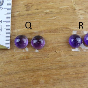 May include: Two pairs of polished, round, deep purple amethyst cabochons, labeled "Q" and "R", are displayed on a wooden surface. A ruler is positioned to the left, indicating size. These gemstones are ideal for jewelry making.
