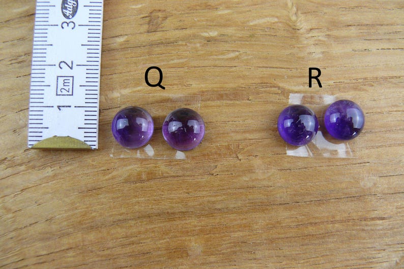 May include: Two pairs of polished, round, deep purple amethyst cabochons, labeled "Q" and "R", are displayed on a wooden surface. A ruler is positioned to the left, indicating size. These gemstones are ideal for jewelry making.
