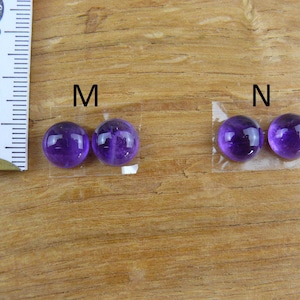 May include: Two pairs of polished, round, deep purple gemstones, labeled "M" and "N", are displayed on a wooden surface. A ruler is positioned next to the "M" pair. The gemstones are likely amethyst, used for jewelry or crafts.