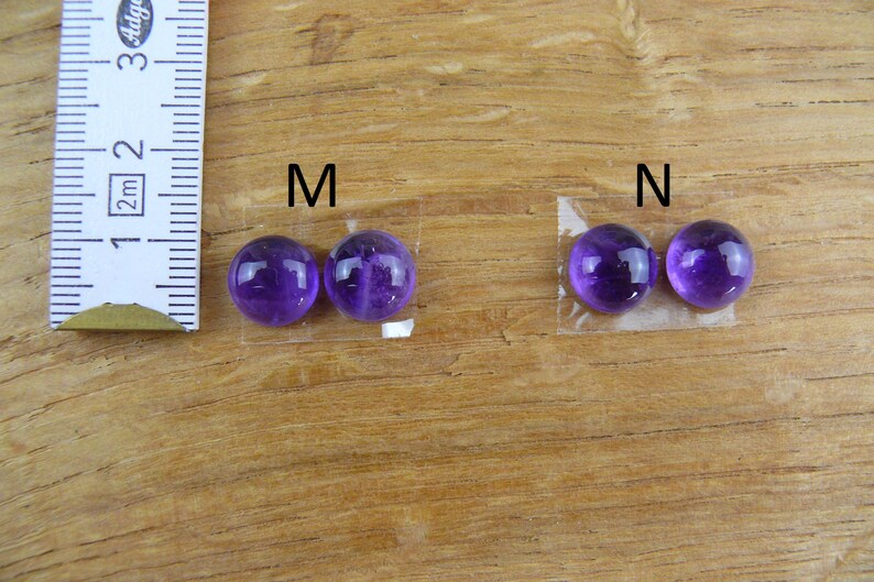 May include: Two pairs of polished, round, deep purple gemstones, labeled "M" and "N", are displayed on a wooden surface. A ruler is positioned next to the "M" pair. The gemstones are likely amethyst, used for jewelry or crafts.