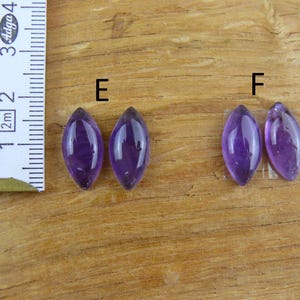 May include: Four polished, marquise-cut amethyst gemstones in varying shades of purple are displayed on a wooden surface. A ruler with metric and imperial measurements is positioned to the left of the gemstones. The gemstones are labeled with the letters "E" and "F".