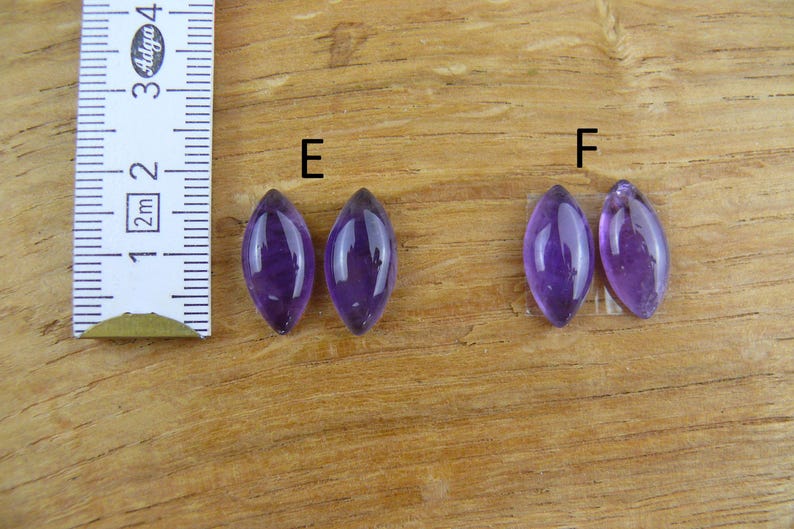 May include: Four polished, marquise-cut amethyst gemstones in varying shades of purple are displayed on a wooden surface. A ruler with metric and imperial measurements is positioned to the left of the gemstones. The gemstones are labeled with the letters "E" and "F".