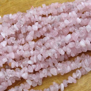 Pode incluir: Close-up de fios de contas de lascas de quartzo rosa claro. As contas são pequenas, de formato irregular e têm uma aparência ligeiramente translúcida. As contas são enfiadas, criando uma superfície fluida e texturizada.