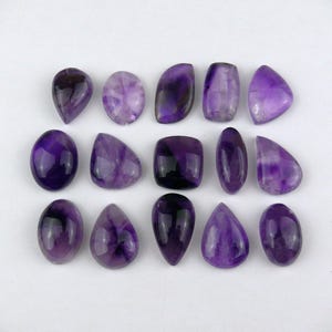 Könnte beinhalten: Eine Sammlung von fünfzehn polierten Amethyst-Edelsteinen, die in drei Reihen angeordnet sind. Die Steine haben verschiedene Formen, darunter Tropfen, Ovale und Rechtecke, und zeigen tiefe violette Farbtöne mit natürlichen Farbvariationen.