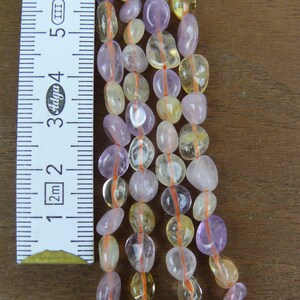 May include: A strand of gemstone beads in shades of purple and yellow. The beads are oval shaped and have a smooth, polished finish. The beads are strung on a thin, orange thread. A ruler is visible in the background, showing the size of the beads.