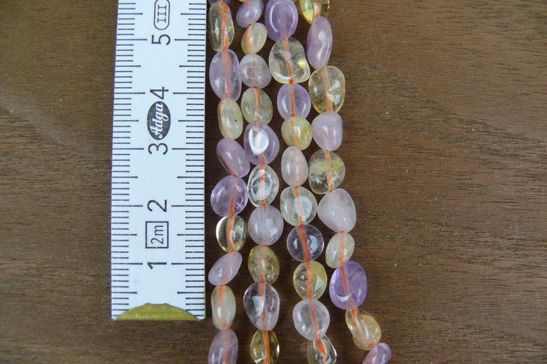 May include: A strand of gemstone beads in shades of purple and yellow. The beads are oval shaped and have a smooth, polished finish. The beads are strung on a thin, orange thread. A ruler is visible in the background, showing the size of the beads.
