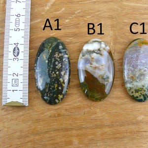 Op de afbeelding: Drie ovaalvormige gepolijste edelstenen met de labels A1, B1 en C1, geplaatst naast een wit meetlint dat een lengte van 7 centimeter aangeeft. De A1-steen is donkergroen met gele stippen, B1 is wit, bruin en groen, en C1 is paars en groen.