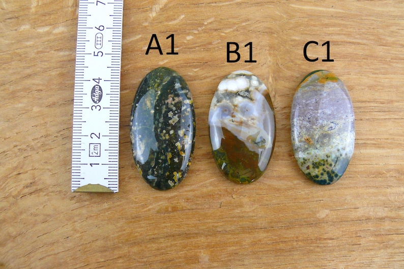 Op de afbeelding: Drie ovaalvormige gepolijste edelstenen met de labels A1, B1 en C1, geplaatst naast een wit meetlint dat een lengte van 7 centimeter aangeeft. De A1-steen is donkergroen met gele stippen, B1 is wit, bruin en groen, en C1 is paars en groen.