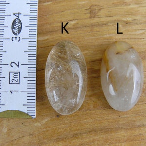 May include: Two oval-shaped clear quartz stones with gold inclusions. The stones are labeled "K" and "L". A ruler is in the background.
