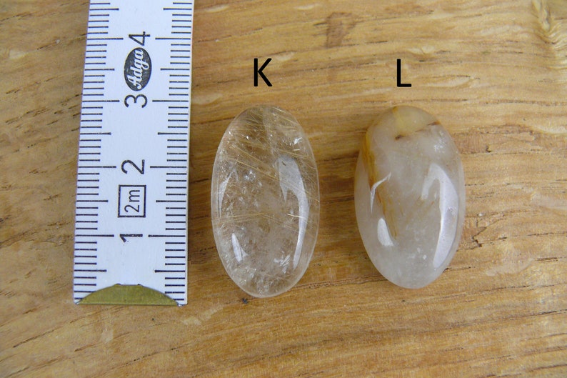 May include: Two oval-shaped clear quartz stones with gold inclusions. The stones are labeled "K" and "L". A ruler is in the background.