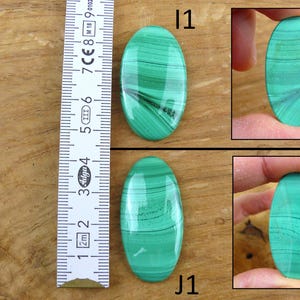 May include: Two oval-shaped malachite gemstones with green and black banding. The gemstones are displayed with a ruler, showing measurements in centimeters. The gemstones are labeled "I1" and "J1".