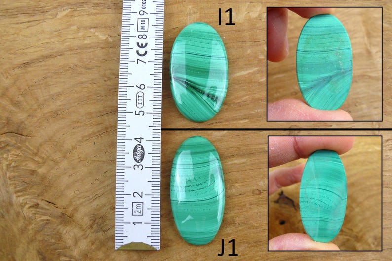 May include: Two oval-shaped malachite gemstones with green and black banding. The gemstones are displayed with a ruler, showing measurements in centimeters. The gemstones are labeled "I1" and "J1".