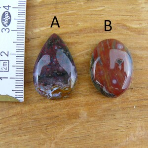 May include: Two polished cabochon gemstones labeled A and B, positioned next to a white ruler showing metric measurements. Gemstone A is teardrop-shaped with deep red, purple, and white inclusions. Gemstone B is oval-shaped with red and white tones.