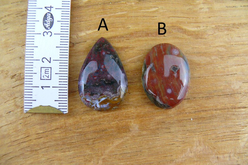 May include: Two polished cabochon gemstones labeled A and B, positioned next to a white ruler showing metric measurements. Gemstone A is teardrop-shaped with deep red, purple, and white inclusions. Gemstone B is oval-shaped with red and white tones.