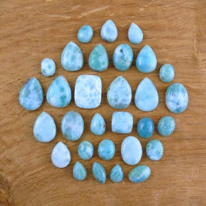 May include: A collection of polished larimar gemstones in various shapes, including teardrop, oval, and rectangular. The stones display a range of blue and turquoise hues with white marbling, set against a wooden surface.