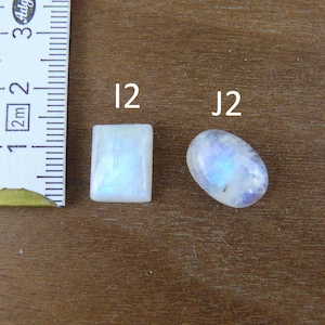 May include: Two moonstone cabochons labeled I2 and J2, with a rectangular stone and an oval stone, both displaying a blue adularescence. A white measuring tape is positioned to the left, showing a scale in centimeters for size reference.