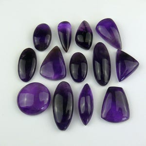 May include: Assortment of 14 amethyst cabochons in various shapes and sizes. The cabochons are a deep purple color and have a smooth, polished finish.