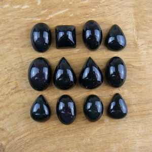 Peut inclure: Une sélection de douze pierres précieuses noires polies de différentes formes, dont ovales, carrées et en forme de goutte. Les pierres sont disposées sur une surface en bois brun clair, reflétant la lumière et mettant en valeur leur finition brillante.