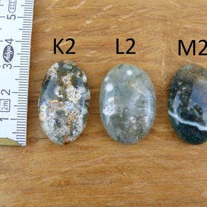 Op de afbeelding: Drie ovaalvormige gepolijste edelstenen met de labels K2, L2 en M2, geplaatst naast een witte liniaal met metingen in centimeters. De stenen hebben gevlekte patronen in tinten donkergroen, grijs, bruin en wit met duidelijke insluitingen.