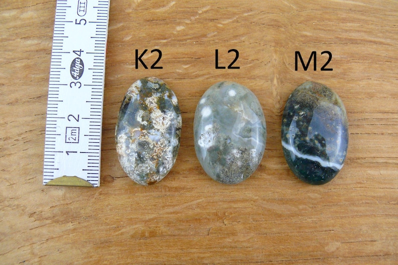 Op de afbeelding: Drie ovaalvormige gepolijste edelstenen met de labels K2, L2 en M2, geplaatst naast een witte liniaal met metingen in centimeters. De stenen hebben gevlekte patronen in tinten donkergroen, grijs, bruin en wit met duidelijke insluitingen.