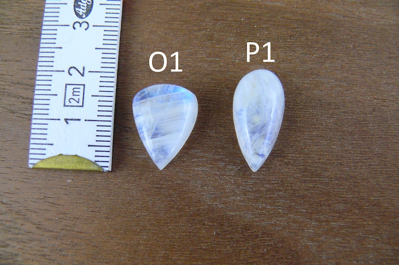 May include: Two pear-shaped moonstone cabochons labeled O1 and P1, positioned next to a white measuring tape showing metric units. The stones are translucent white with blue adularescence, displaying smooth, polished surfaces and pointed ends.