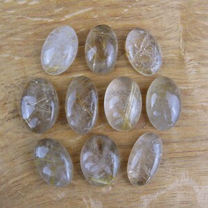 May include: Ten oval-shaped clear quartz stones with gold inclusions. The stones are arranged in a grid pattern on a wooden surface.