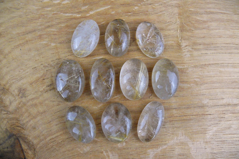 May include: Ten oval-shaped clear quartz stones with gold inclusions. The stones are arranged in a grid pattern on a wooden surface.
