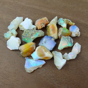 Könnte beinhalten: Eine Sammlung roher Opal-Edelsteine in verschiedenen Farben, darunter Weiß, Orange, Blau und Grün. Die Opale sind grob geschnitten und haben ein natürliches, unpoliertes Aussehen. Die Edelsteine sind auf einer braunen Holzoberfläche angeordnet.