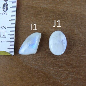 May include: Two polished moonstone cabochons labeled I1 and J1, positioned next to a white measuring tape showing metric units. The I1 stone is a triangular shape with blue adularescence, while the J1 stone is an oval shape with a white hue.