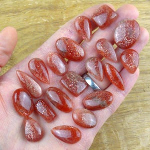 May include: A selection of polished, reddish-orange sunstone gemstones in diverse shapes, including oval, teardrop, and rectangular. The stones have a sparkling, glittery appearance, held in a hand. The gemstones are translucent and appear to have inclusions.