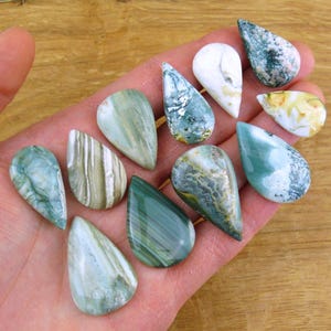 Può includere: Una collezione di undici pietre preziose levigate a forma di goccia in varie tonalità di verde, bianco e crema. Le pietre presentano motivi diversi, tra cui bande vorticose, inclusioni simili a muschio e superfici opache con finitura liscia.
