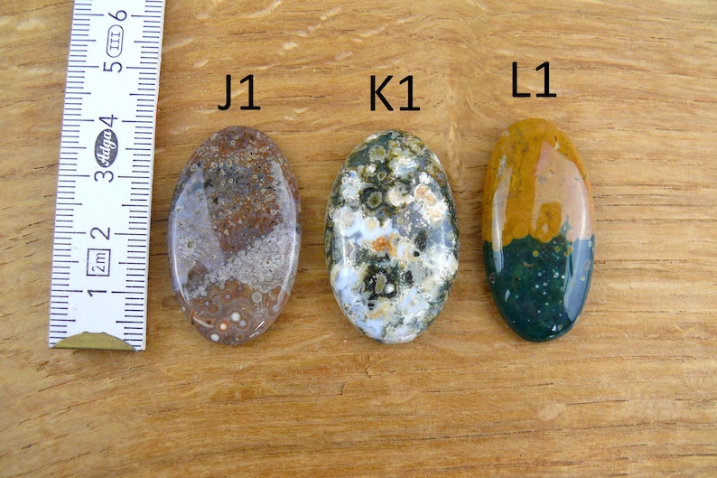 Op de afbeelding: Drie ovaalvormige gepolijste edelstenen met de labels J1, K1 en L1. J1 is bruin met gespikkelde patronen, K1 is groen en wit met gevlekte stippen en L1 is verdeeld tussen geel en donkergroen. Een liniaal toont een schaalverdeling in cm.