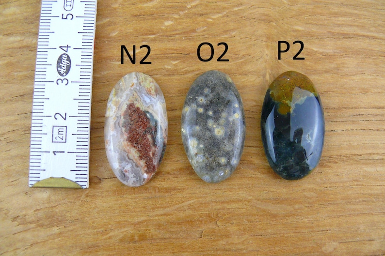 Op de afbeelding: Drie ovaalvormige gepolijste edelstenen met de labels N2, O2 en P2. De eerste is cr&egrave;me en bruin, de tweede is donkergrijs met gele stippen en de derde is donkerblauw en bruin. Een witte liniaal toont een lengte van ongeveer 4 cm.