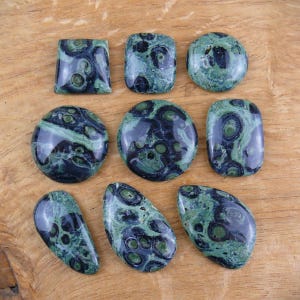 Peut inclure: Neuf pierres précieuses polies de différentes formes, dont carrées, ovales et rondes. Les pierres présentent un motif vert foncé et noir, avec une finition brillante. Les pierres sont exposées sur une surface en bois brun clair.