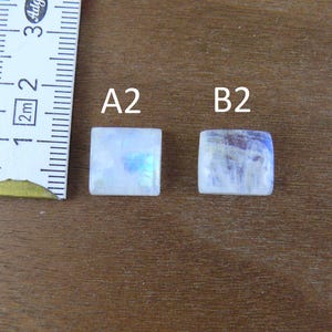 May include: Two square moonstone cabochons labeled A2 and B2, positioned next to a white measuring tape showing metric units. The stones exhibit a translucent white appearance with subtle blue iridescence and smooth, flat surfaces.