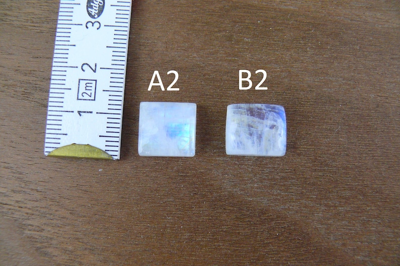 May include: Two square moonstone cabochons labeled A2 and B2, positioned next to a white measuring tape showing metric units. The stones exhibit a translucent white appearance with subtle blue iridescence and smooth, flat surfaces.