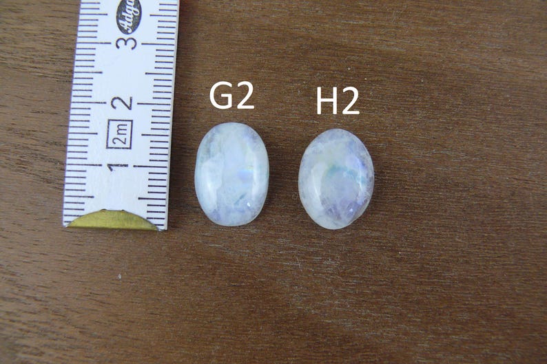 May include: Two oval-shaped, translucent white moonstones labeled G2 and H2, positioned next to a white measuring tape showing metric increments. The stones exhibit a subtle blue iridescent sheen and a smooth, polished surface finish.