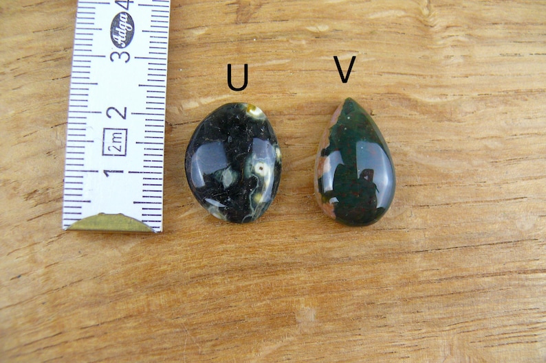 May include: Two polished gemstones labeled U and V next to a ruler. Gemstone U is oval, dark green with white spots. Gemstone V is teardrop-shaped, dark green with a tan section. The ruler shows a scale in centimeters for size reference.