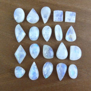May include: A collection of twenty polished moonstone cabochons arranged in four rows. The gemstones feature various shapes including ovals, teardrops, squares, and diamonds, all displaying a translucent white appearance with subtle blue adularescence.