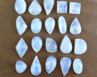 May include: A collection of twenty polished moonstone cabochons arranged in four rows. The gemstones feature various shapes including ovals, teardrops, squares, and diamonds, all displaying a translucent white appearance with subtle blue adularescence.