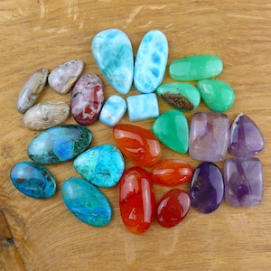 Puede incluir: Una colección de piedras preciosas pulidas de diversas formas y tamaños, que incluye larimar azul vibrante, cornalina roja intensa, crisoprasa verde brillante, amatista morada y piedras de ágata con patrones, todas con superficies lisas y brillantes.