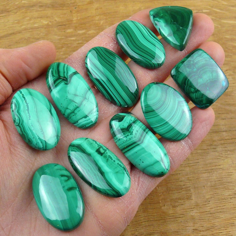 May include: A collection of polished malachite gemstones in various shapes, including ovals and a triangle. The stones display a vibrant green color with distinctive swirling patterns. The gemstones are held in a hand, showcasing their natural beauty.